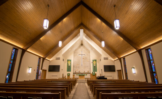 The center aisle lined with pews leads to the church platform with illuminated cross above the alter. At the crux of the high ceiling, a white line array points towards the congregation.
