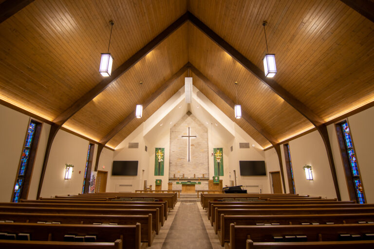 The center aisle lined with pews leads to the church platform with illuminated cross above the alter. At the crux of the high ceiling, a white line array points towards the congregation.