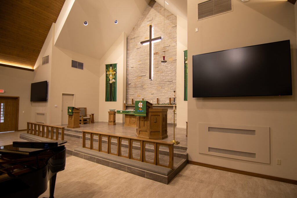 A side view of the platform in the sanctuary of Hope Lutheran Church. An illuminated cross on a stone wall, green tapestries, prayer railing, pulpit, alter, and worship system with large video displays.
