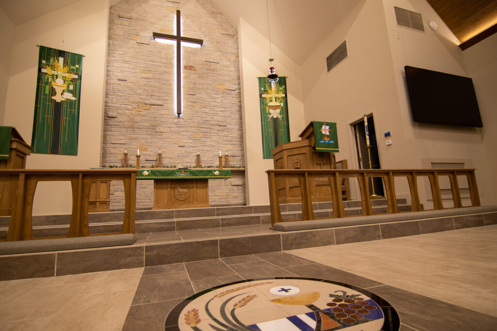 Close up of the platform of Hope Lutheran Church. An illuminated cross on a stone wall, green tapestries, prayer railing, pulpit, alter, and worship system with video and microphones.