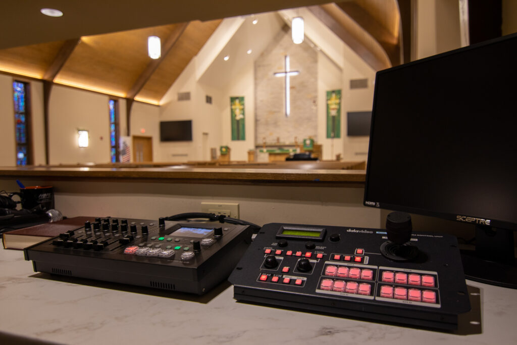A close up of the video portion of the worship system. A joystick controller and computer monitor on the right, a video mixer with lots of knobs and buttons on the left. The front platform can be seen in the background, blurred.