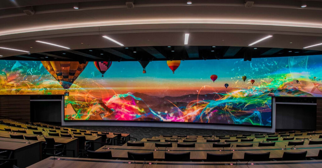 A massive direct view LED video wall covering the entire front wall of a lecture hall. The image displayed is a colorful array of hot air balloons over a dessert sunset.