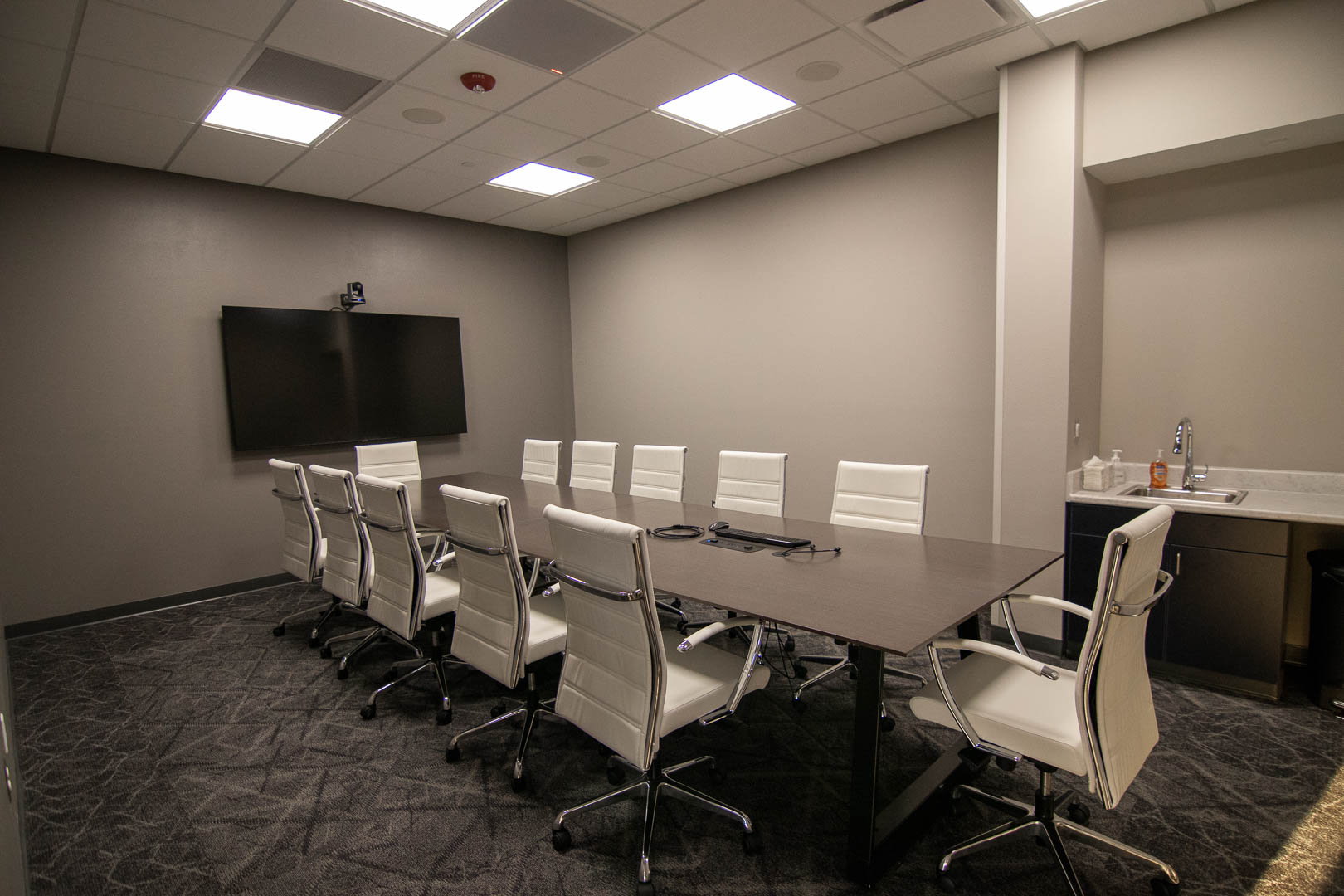 A conference room connected to the larger event space. 10 white office chairs surround a conference table with a large flat panel display at one end. A conferencing camera sits above the TV. A microphone is cleverly disguised as a ceiling tile but the LED mute is a dead giveaway.